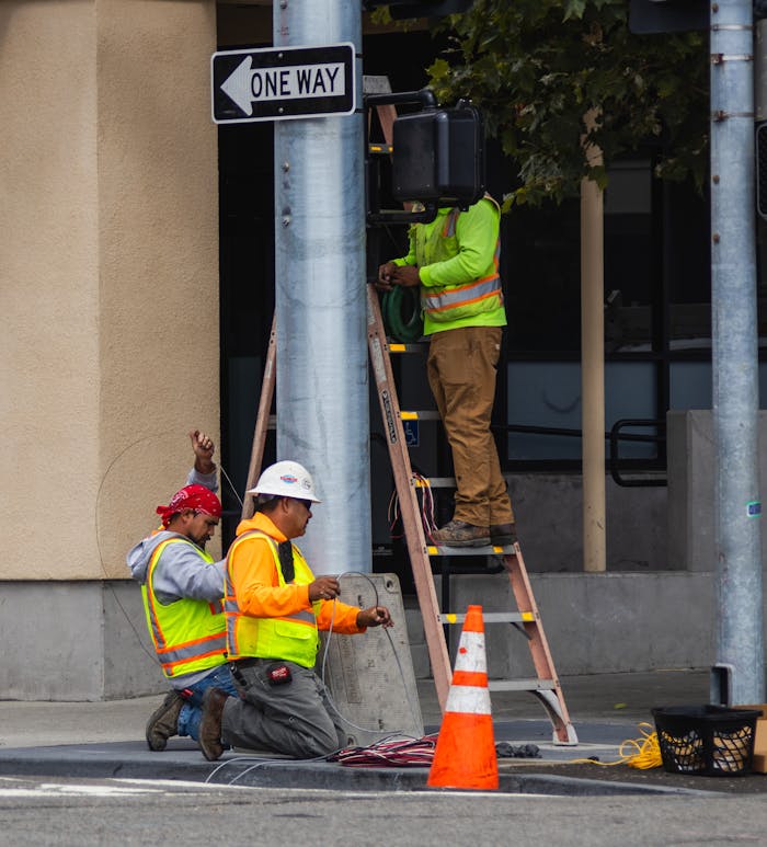 City workers in reflective gear maintain and repair a traffic signal on a bustling urban street.